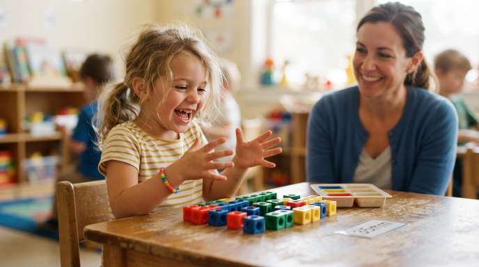 Patient teacher smiling at a young child sitting at a wooden table with colorful math counting blocks, encouraging trial and error in kids